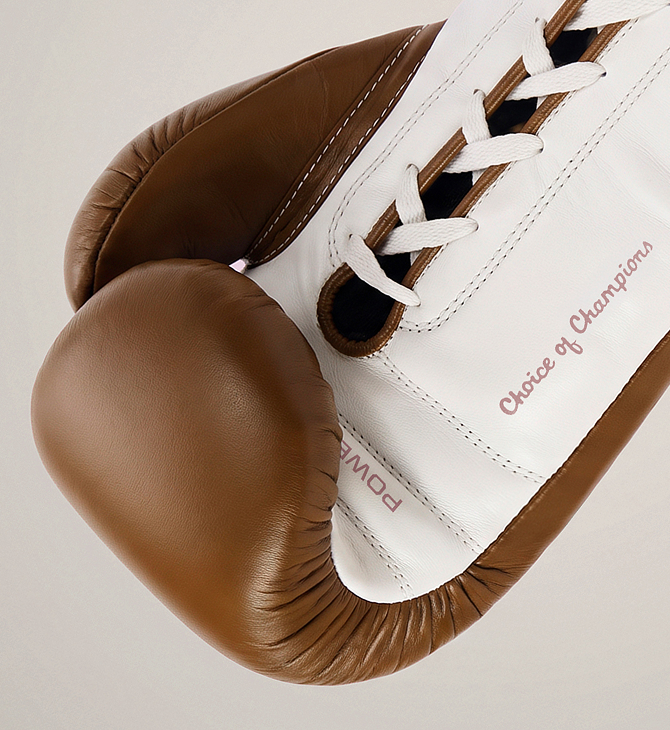 Close-up of a boxing glove in brown and white leather with white laces and the words 'Choice of Champions' printed on the side, photographed on a light neutral background.