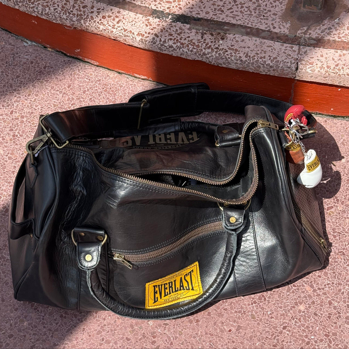 A leather Everlast duffel bag with a yellow brand patch on the front, resting on a terrazzo-patterned floor next to a metal railing. Brown red white pink black