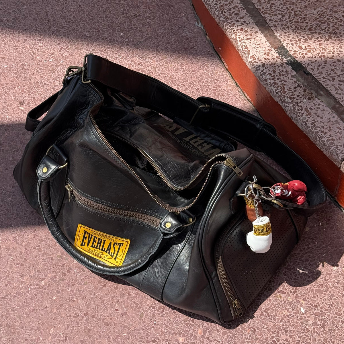 A leather Everlast duffel bag lying on a paved surface outdoors, with a 1910 Everlast logo patch on the front pocket and a keychain attached. Brown red white pink black